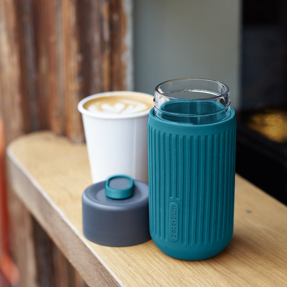 BLACK+BLUM blue glass travel mug with open lid and coffee, outdoor ocean scene, stylish reusable Kaffeebecher for on-the-go.